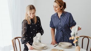 A mother and daughter setting the table with plates, cutlery, and flowers in a bright room.
